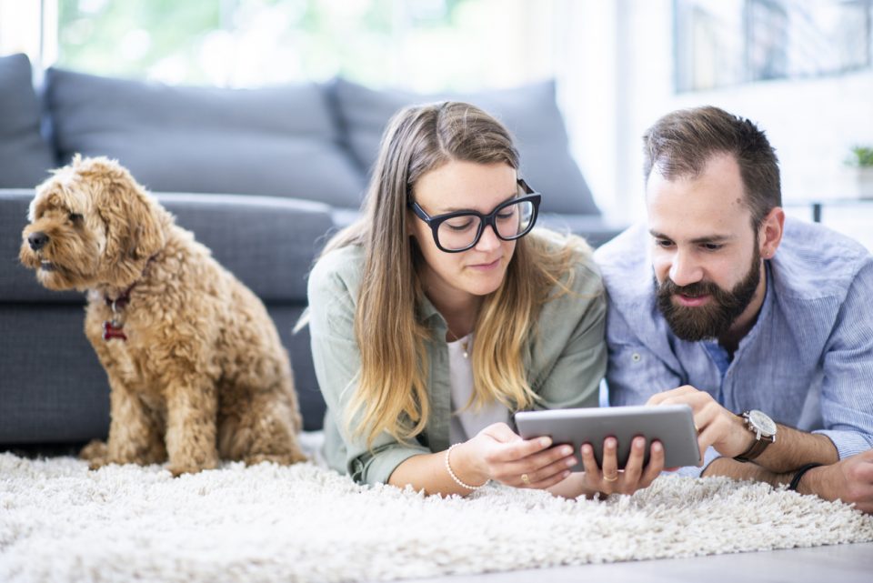 A caucasian couple lies on their living room rug, with their dog in the background, looking at a tablet.