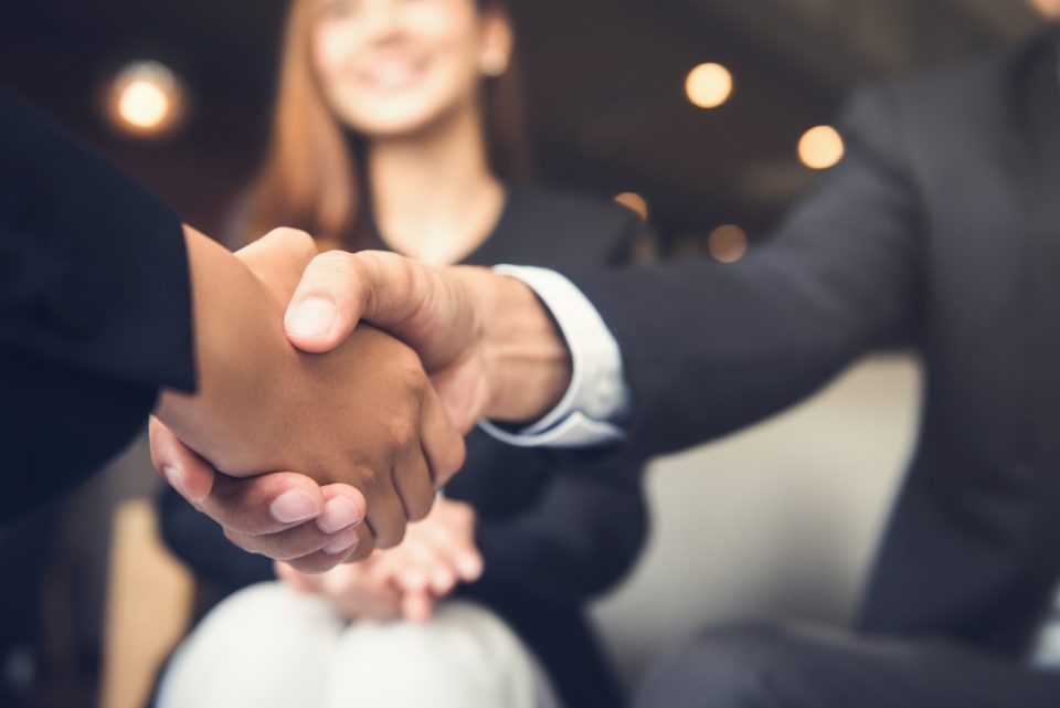 Businessmen shaking hands, wearing suits while female colleague smiles in background