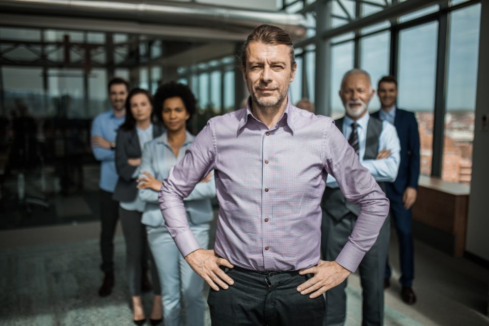Confident mid adult businessman standing in front of his team with hands on hips and looking at camera.