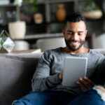 Cheerful latin american man relaxing on couch while looking at social media on tablet smiling