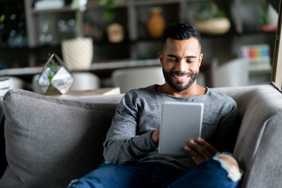 Cheerful latin american man relaxing on couch while looking at social media on tablet smiling